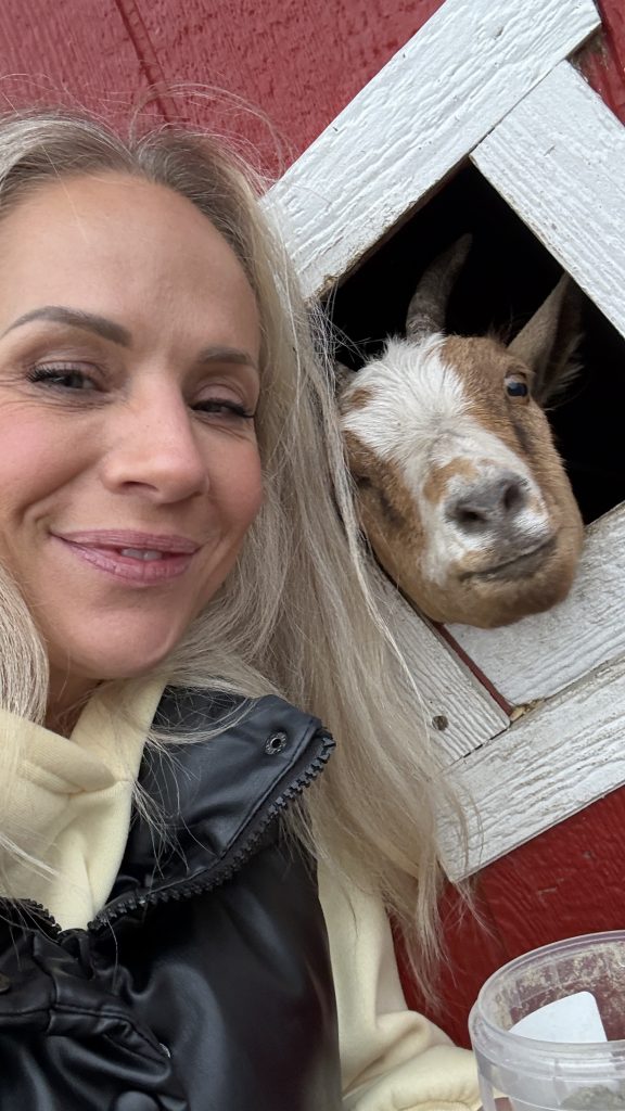 Carrie Meir taking a selfie with a curious goat poking its head out of a red barn window at Kraay Family Farm