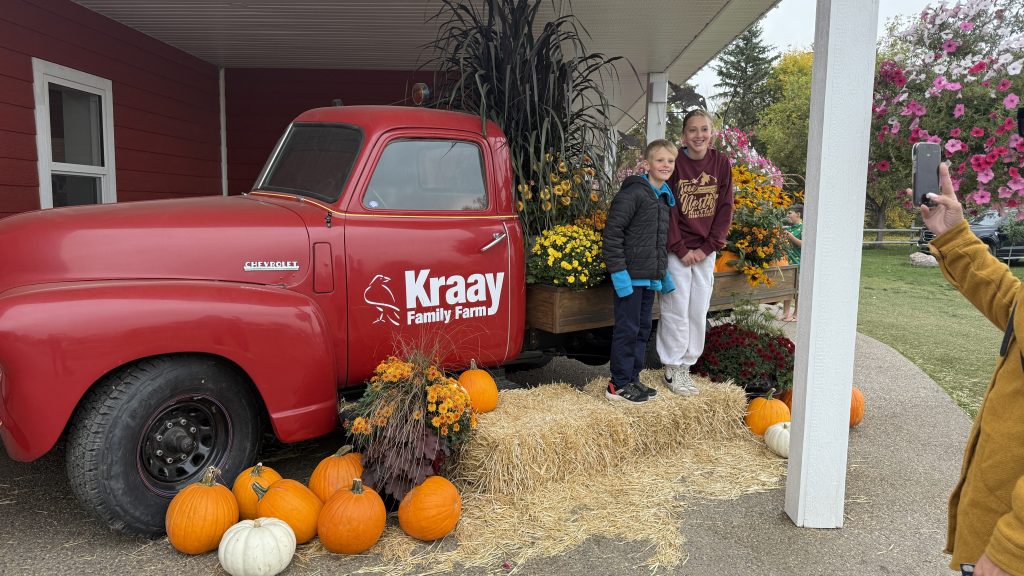Two kids posing beside the red vintage Kraay Family Farm truck decorated with pumpkins and fall mums